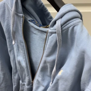 Ljusblå hoodie med dragkedja från Polo Ralph Lauren - Ljusblå hoodie från Polo Ralph Lauren med dragkedja framtill och vit broderad logga på bröstet. Tröjan har huva med dragsko och mjukt material på insidan. Perfekt för en avslappnad stil.