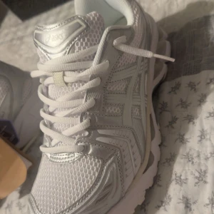 Vita ASICS GEL-Kayano 14 sneakers - Säljer ett par fräscha vita ASICS GEL-Kayano 14 jjjjound Collab sneakers med silvriga detaljer. Skorna har mesh- och syntetmaterial, klassisk snörning och en chunky sula. Perfekta för dig som gillar sportig stil och vill ha bekväma skor till vardagen.