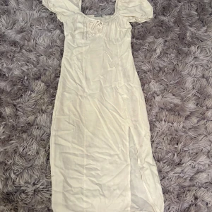 Vit långklänning med puffärmar från DeFacto - Superfin vit långklänning från DeFacto med puffiga kortärmar och söt knytdetalj vid bysten. Klänningen har en slits på sidan och är gjord i ett lätt material, perfekt för varma dagar.