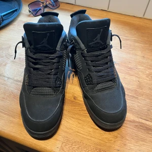 Jordan 4 blackcats - Säljer ett par helsvarta Air Jordan sneakers med klassisk siluett och snörning. Skorna har en robust sula och detaljer i mesh och mocka. Perfekta för dig som gillar streetwear och vill ha en stilren look.