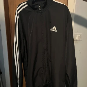 Svart träningsjacka från Adidas - Svart träningsjacka från Adidas med klassiska vita ränder längs ärmarna och vit Adidas-logga på bröstet. Jackan har dragkedja framtill och lång ärm. Perfekt för sportiga tillfällen eller som en snygg streetwear-detalj.