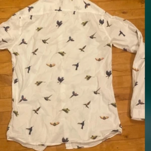 Vit skjorta med fågelmönster från Lindbergh - Säljer en vit skjorta från Lindbergh med färgglatt fågelmönster över hela plagget. Skjortan har långa ärmar och klassisk krage. Perfekt för dig som vill sticka ut med en unik och stilren look.