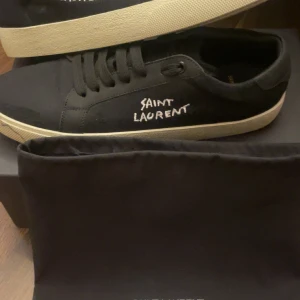 Svarta sneakers från Saint Laurent - Svarta sneakers från Saint Laurent med vit sula och handskriven logga på sidan. Skorna har rund tå, platt sula och svarta snören. Klassisk och stilren design med diskreta detaljer och Saint Laurent Paris-tryck på plösen och sulan.Storlek 41 men passar 42, Box dustbag och kvitto finns, Såklart äkta. 