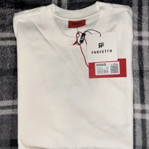 Vit t-shirt från HUGO - Säljer en stilren vit t-shirt från HUGO med diskret broderad logga på bröstet. Klassisk rund halsringning och normal passform. Perfekt basplagg för garderoben. Märkeslapp och taggar från Farfetch sitter kvar.