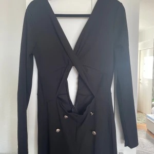 Svart långärmad jumpsuit med djup v-ringning - Säljer en svart långärmad jumpsuit med djup v-ringning och snygga knappar framtill. Jumpsuiten har en åtsittande passform och markerad midja, perfekt för att skapa en stilren look.