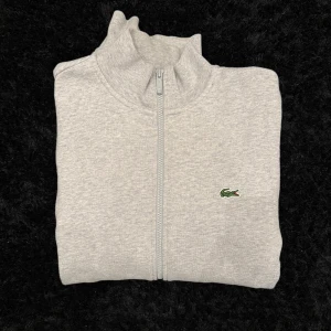 Grå ziptröja från Lacoste - Säljer en stilren grå ziptröja från Lacoste med hög krage och dragkedja framtill. Tröjan har det klassiska gröna krokodilmärket på bröstet och långa ärmar. Perfekt för en avslappnad och sportig look.