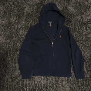 Mörkblå hoodie från Polo Ralph Lauren - Säljer en mörkblå hoodie från Polo Ralph Lauren med dragkedja och huva. Tröjan har en röd broderad logga på bröstet och två fickor framtill. Perfekt för en avslappnad stil.