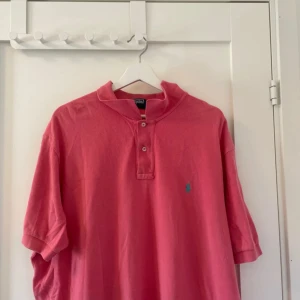 Rosa pikétröja från Polo Ralph Lauren - Säljer en klassisk rosa pikétröja från Polo Ralph Lauren med turkos broderad logga på bröstet. Tröjan har korta ärmar, knappar vid halsen och är i bomull. Perfekt för en avslappnad och stilren look. Endast 249kr pris ej hugget i sten