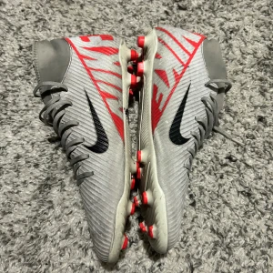 Nike Mercurial fotbollsskor i grått och rött - Säljer ett par Nike Mercurial fotbollsskor i grått med röda och svarta detaljer. Skorna har snörning, högre skaft och dobbar för spel på gräs. Swoosh-logga på sidan och röd text längs hälen. Perfekta för dig som vill sticka ut på planen.