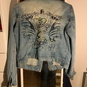 Jeansjacka med broderad rygg - Säljer en klassisk blå jeansjacka med unikt broderi på ryggen som föreställer en drake, blommor och dödskalle. Jackan har knappar framtill och två bröstfickor. Perfekt för dig som vill sticka ut med en cool detalj.