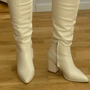 Beiga höga boots med klack - Säljer ett par beiga höga boots med bred klack och spetsig tå. Skorna har dragkedja på insidan och är tillverkade i syntetmaterial. Perfekta för att lyfta din outfit med en trendig touch. Bilder på defekter syns på bilderna. Vet inte vart dom är ifrån. 🩷står ingen storlek men passar mig som annars har strl 38