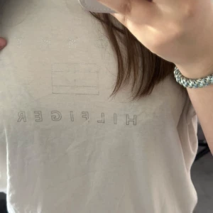 Vit t-shirt från Tommy Hilfiger - Vit t-shirt från Tommy Hilfiger med diskret logga och broderad text framtill. Klassisk passform och rund halsringning. Perfekt till jeans eller shorts för en clean look.