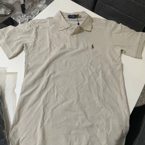 Beige tshirt från Polo Ralph Lauren - Säljer en beige pikétröja från Polo Ralph Lauren med klassisk krage och två knappar framtill. Tröjan har broderad logga på bröstet och är i en slim fit-modell. Perfekt för en stilren och avslappnad look. Den är inte normal i storlek står S men skulle mer säga den passar dig i M 