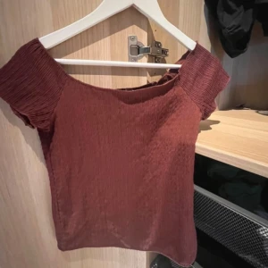 Rostbrun croppad topp från 157 - Säljer en rostbrun croppad topp från 157 med korta ärmar och smockad struktur. Toppen har en bred halsringning och är tillverkad i ett stretchigt material som sitter snyggt på kroppen.