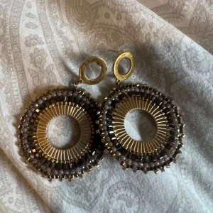 Stora runda örhängen i guld och brunt - Säljer ett par stora, runda örhängen i guld med bruna och guldfärgade pärldetaljer. Örhängena har en öppen cirkelform och är dekorerade med små pärlor runt kanten för en bohemisk och unik look. Använda 1-3 gånger säljer pga att det inte riktigt är min smak😍