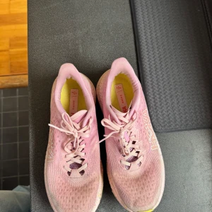 Rosa löparskor från Hoka - Säljer ett par rosa Hoka löparskor med vit logga på sidan och ljusgul innersula. Skorna har snörning och en tjock, mönstrad sula i vitt och gult. Perfekta för löpning eller promenader.