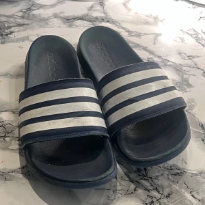 Adidas badsandaler i mörkblått - Mörkblå Adidas sandaler med klassiska vita ränder över foten. Perfekta för stranden eller poolen, tillverkade i syntetmaterial för att tåla vatten. Enkel och sportig design med platt sula och öppen tå.