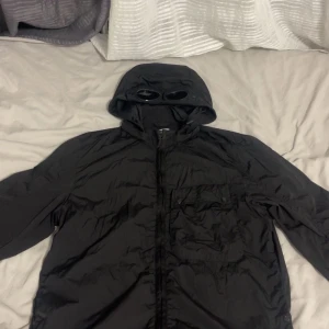 Svart Goggle Jacket från C.P. Company - Säljer en svart Goggle Jacket från C.P. Company med ikoniska glasögon i huvan. Jackan är tillverkad i återvunnen nylon och har dragkedja framtill. Perfekt för dig som gillar streetwear och unika detaljer.