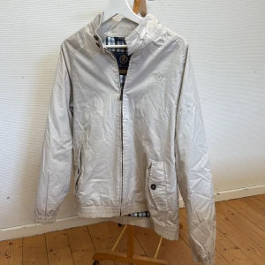 Vit vindjacka från Henri Lloyd - Snygg beige vindjacka från Henri Lloyd med dragkedja och broderad logga på bröstet. Jackan har långärmade ärmar, rutigt innerfoder och praktiska fickor med knappstängning. Perfekt för vår och höst. Sitter som M.
