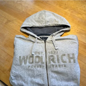 Grå Woolrich Zip Hoodie - Huvtröja med dragkedja från Woolrich med broderat tryck, i fint skick. Storlek: US/M - EU/L. Passar bra på mig, jag har storlek M i EU storlekar. Tveka inte på att höra av er vid frågor! 😄
