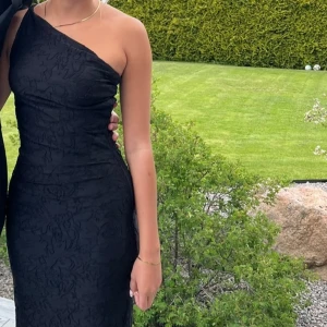 Svart oneshoulder långklänning - Säljer en elegant svart långklänning med oneshoulder-design och diskret mönster i tyget. Klänningen har en figurnära passform och är perfekt för speciella tillfällen som bal. Köpt från Boohoo men omsydd.