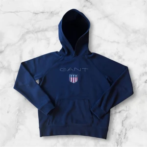 Marinblå hoodie från GANT - Säljer en marinblå hoodie från GANT med broderad logga och sköld på bröstet. Tröjan har huva, magficka och långa ärmar. Perfekt för en avslappnad stil och enkel att matcha med jeans eller joggers.