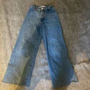 Blå wide leg jeans från Zara - Säljer ett par blå jeans från Zara i wide leg-modell med råa kanter nertill. Byxorna har hög midja, klassiska fickor bak och knappstängning framtill. Perfekta för en avslappnad och trendig look.