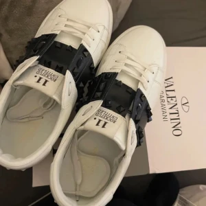 Valentino sneakers med svarta nitar - Säljer ett par vita Valentino sneakers med bred svart rem över foten dekorerad med svarta nitar. Skorna har rund tå, platt sula och klassisk snörning. Märkeslogga på plösen och detaljerade sömmar. Lite röd fläck vid de svarta som ej syns mycket alls skriv privat för mer bilder