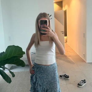 Blå blommig omlottkjol med volang - Säljer en blå omlottkjol med vitt blommönster och volangkant. Kjolen har knytband i midjan och är i en lätt och luftig kvalitet, perfekt för varmare dagar. Kjolen är kort och har en feminin och somrig stil.