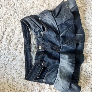 Jeanskjol med volang från D&G - Säljer en snygg mörkblå jeanskjol från D&G med volang nertill. Kjolen har klassiska jeansfickor fram och bak samt knappar och dragkedja framtill. Perfekt för dig som gillar denim och vill ha något unikt i garderoben. Midjemått 35 men stretchig o längden är 31!!