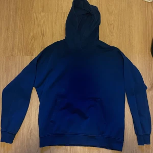 Blå hoodie från Lindex - En enkel och stilren blå hoodie från Lindex i storlek L. Tröjan har huva, lång ärm och en klassisk magficka framtill. Tillverkad av återvunnen och ekologisk bomullsblandning, perfekt för en avslappnad look.