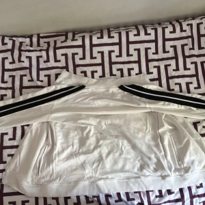 Vit Palm Angels M - Snygg vit zipp upp hoodie  från Palm Angels med svarta ränder längs ärmarna och dragkedja framtill. Jackan har broderad logga på bröstet och två fickor med dragkedja. Tillverkad i polyester för en sportig vibe.