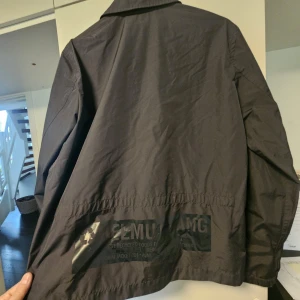 Svart vindjacka från Helmut Lang - Svart vindjacka från Helmut Lang med klassisk krage och tryckknappar framtill. Jackan har diskret logga på bröstet och detaljerad text på ryggen. Tillverkad i 100% återvunnen polyester från havsplast, vilket gör den både snygg och miljövänlig. Nypris +4000:-