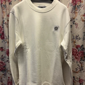 Vit sweatshirt från Stone Island - Säljer en stilren vit sweatshirt från Stone Island med klassisk rund hals och logotyp-patch på bröstet. Tröjan har långa ärmar och är tillverkad i mjuk bomull, perfekt för en clean och enkel look.