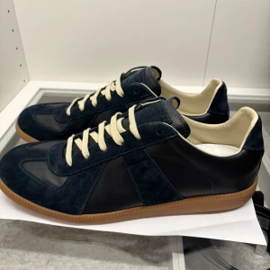 Marinblå sneakers med mockadetaljer - Snygga marinblå sneakers från Maison Margiela med ovandel i mocka och skinn, beige snörning och gummisula. Skorna har en klassisk siluett och kontrasterande ljus insida. Perfekta för dig som gillar stilrena och trendiga sneakers.