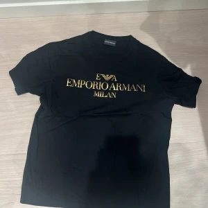 Svart t-shirt från Emporio Armani - Snygg svart t-shirt från Emporio Armani med guldig logga och text på bröstet. Klassisk rund halsringning och korta ärmar. Tillverkad i mjuk bomull för en skön känsla hela dagen. Storlek M men passar S. Pris kan diskuteras vid snabb affär.