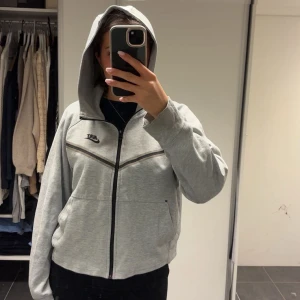 Nike Tech hoodie - Säljer en grå zip-hoodie/ Nike Tech från Nike med svart logga på bröstet och svarta detaljer längs dragkedjan och bröstet. Jackan har huva, långa ärmar och två fickor framtill. Perfekt för en sportig och avslappnad stil. Lågt pris på grund av deffekterna på 2 sista sidorna, de syns däremot ej vid använding!!