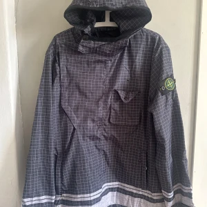Grå vindjacka med reflex från Stone Island - Säljer en grå vindjacka från Stone Island med vitt reflex rutmönster och huva. Jackan har en bröstficka, klassisk logotyp på ärmen och tryckt logga på ryggen. Perfekt för dig som gillar streetwear och snygga detaljer.