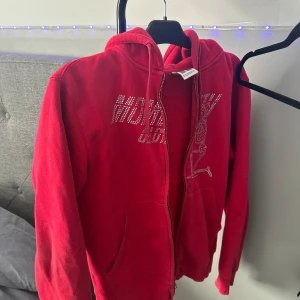 Röd hoodie med dragkedja från Russell - Säljer en röd hoodie från Russell i storlek XS. Tröjan har dragkedja framtill, huva och fickor. Dekorerad med glittrig text på bröstet. Perfekt för dig som gillar en färgstark och bekväm stil.