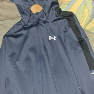 Blå träningsjacka med huva från Under Armour - Snygg blå träningsjacka från Under Armour med svart detalj längs ärmen och vit logga på bröstet. Jackan har huva och dragkedja framtill, perfekt för aktiva dagar.