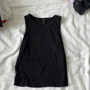 Svart ribbad linnetopp från Gina Tricot - En svart ribbad linnetopp från Gina Tricot med klassisk rund halsringning och ärmlös design. Perfekt basic-plagg att matcha med allt. Mjuk och stretchig bomullskänsla som sitter snyggt och bekvämt.