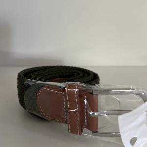 Olivgrönt flätat bälte - Snyggt olivgrönt bälte med flätad design och bruna skinn-detaljer vid spännet. Spännet är i silverfärgad metall och bältet har en klassisk look som passar till många outfits. Passar bra till linnebyxor och mycket mer!