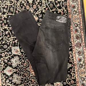 Lee Luke 30/32 slitningar  - Säljer ett par svarta Lee Luke jeans med diskreta slitningar framtill. Jeansen har fem fickor, normal passform och klassisk Lee-lapp bak i midjan. Perfekta för dig som gillar en stilren look med lite edge.