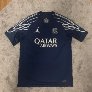 Blå PSG fotbollströja  - Mörkblå PSG fotbollströja i samarbete med Jordan. Tröjan har vita detaljer, vingmönster på axlarna, PSG-logga och Jordan-logga på bröstet samt Qatar Airways som sponsor. Namn och nummer på ryggen: João Neves 87.