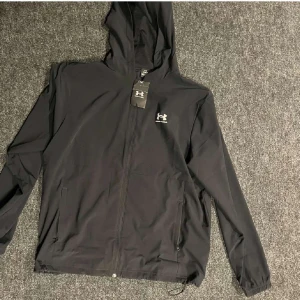 Svart vindjacka från Under Armour - Säljer en svart vindjacka från Under Armour med huva och dragkedja framtill. Jackan har två sidofickor med dragkedja och elastiska muddar vid ärmsluten. Perfekt för blåsiga dagar och sportiga aktiviteter.