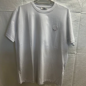 Vit Moncler t-shirt med broderad logga - Snygg vit t-shirt från Moncler i bomull med klassisk rund hals och korta ärmar. På bröstet finns en broderad Moncler-logga i vitt, rött och blått. På ena ärmen finns även ett diskret broderat Moncler-märke. Perfekt för en clean och stilren look. Obs! Finns i alla storlekar. Skriv önskad storlek efter köp! 