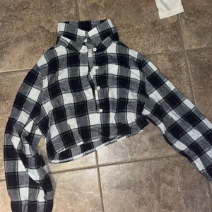 Svartvit rutig croppad skjorta Shein - Croppad skjorta från Shein i svartvitt rutigt mönster. Skjortan har långa ärmar, klassisk krage och knäppning framtill med vita knappar. Perfekt för en chill och trendig look.