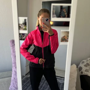 Nike Storm-FIT träningsjacka i rosa och svart - Nike Storm-FIT jacka i starkt rosa och svart med dragkedja framtill och höga fickor med dragkedja. Jackan har lång ärm, hög krage och liten bröstficka med dragkedja. Perfekt för utomhusträning.