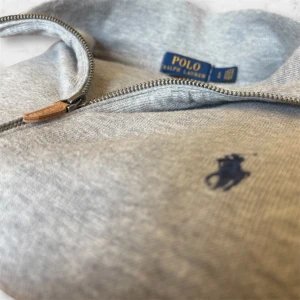 Grå zip-tröja från Polo Ralph Lauren - Snygg grå zip-tröja från Polo Ralph Lauren med klassisk broderad logga på bröstet. Tröjan har hög krage, dragkedja framtill och två fickor. Perfekt för lager-på-lager och enkel att matcha med det mesta.