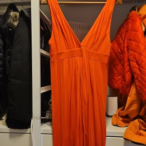 Orange/ rosa  långklänning med v-ringning - Säljer en snygg orange/ rosa långklänning med djup v-ringning och breda axelband. Klänningen har en lös passform och faller fint längs kroppen, perfekt för sommarens alla tillfällen.